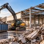 Commercial demolition site with an excavator loading recovered scrap metal into a skip beside a dismantled industrial warehouse